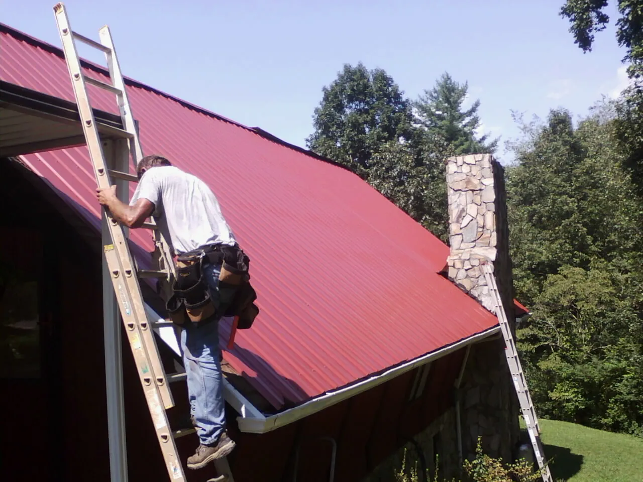 Roofer on a ladder beside a red metal roof and stone chimney — Soffit & Fascia Repair by Roofing Brothers in Wilkes Barre
